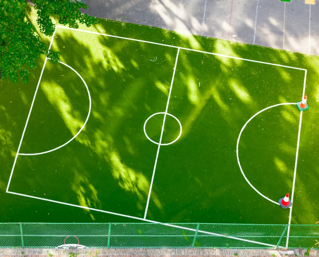 An overhead, bird's-eye view of an empty green artificial turf pitch, ideal for practicing 5 aside football tactics. The pitch features crisp white markings, including a center circle, a halfway line, and two penalty areas, with several orange and white training cones placed on one side. Shadowy silhouettes of nearby trees stretch across the surface, and an asphalt playground area with colorful painted shapes is visible at the top.