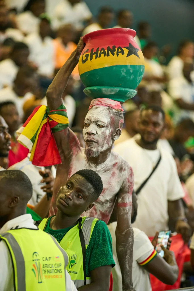A football fan at a crowded stadium holds a clay pot painted in Ghana&rsquo;s national colors&mdash;red, yellow, and green&mdash;with the word &ldquo;GHANA&rdquo; and a black star on it. The fan&rsquo;s face and body are painted, and a Ghana flag is tied around their arm. The black star, a symbol of African freedom and unity, comes from the Ghanaian flag and represents the &ldquo;Black Star of Africa,&rdquo; a sign of independence and leadership in African liberation.