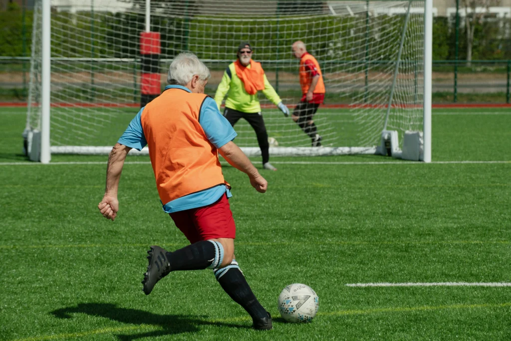 Older men playing a slower-paced game of soccer on a grass pitch, with one player kicking the ball toward the goal while others defend, illustrating an active match of walking football for seniors.