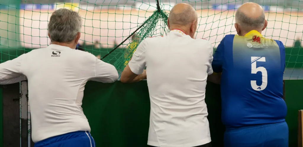 Three older men leaning on a barrier and watching an indoor football pitch through a net, one wearing a blue jersey with the number 5, illustrating what is walking football, a slower-paced version of the sport often played by older adults.