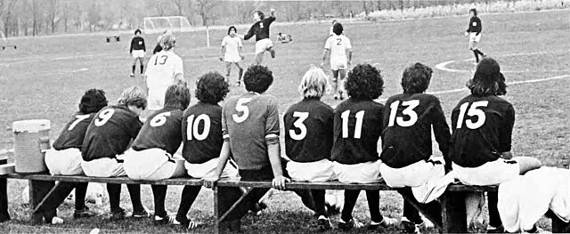 Football team sitting on a bench watching a match on the field, with visible jersey numbers 9, 6, 10, 5, 3, 11, 13, and 15, showing multiple football team numbers lined up across the players&rsquo; backs.