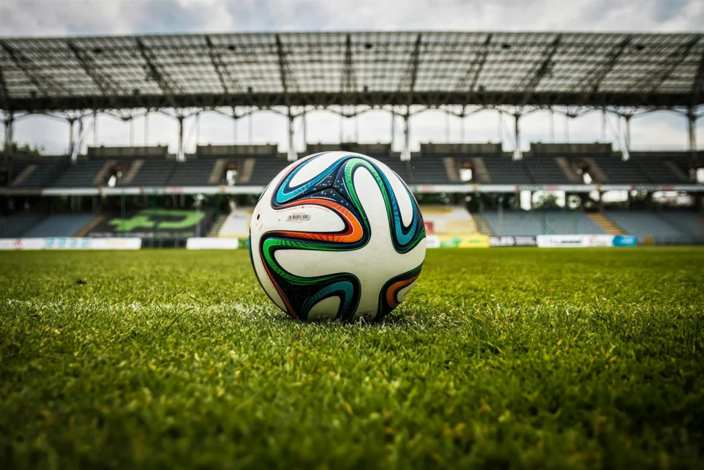 A colorful football sits stationary on a lush green grass field in the center of a large, empty professional stadium. The vast, open space surrounding the ball represents the environment where a player must apply their football iq to perceive tactical information and make accurate decisions under pressure.