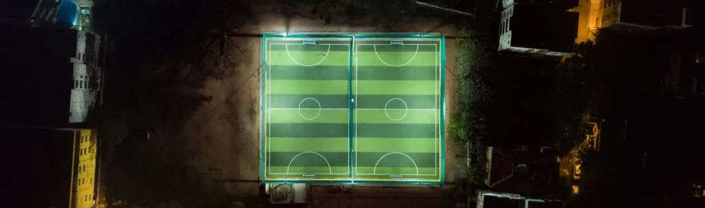 Aerial night view of two football pitches side by side in a city, illustrating the question are football pitches the same size.