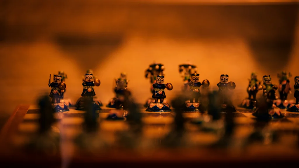 A set of ornate, hand-painted traditional chess pieces arranged on a board under warm, dim lighting. The focus is on the middle row of figures, which resemble small warriors in blue and gold attire. This strategic setup serves as a visual metaphor for football iq, representing the mental chess match and pattern recognition required to anticipate an opponent's next move on the pitch.