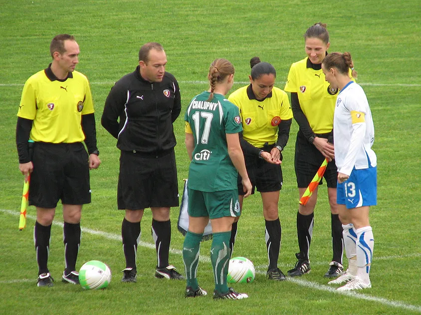 A group of officials and players gather on a grass field for a pre-match football coin toss. A female referee in a yellow jersey looks down at the coin in her hand, surrounded by assistant referees and the two team captains in their respective green and white kits.