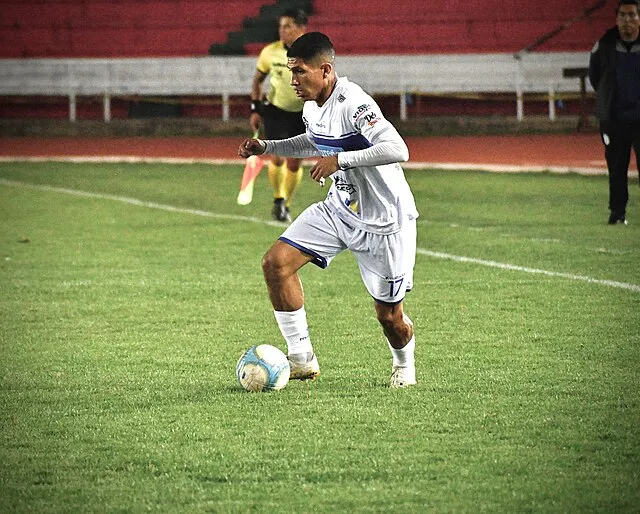 A football player in a white uniform dribbling the ball during a night match on a grass field while a referee and sideline staff stand in the background, illustrating gameplay context for understanding how is possession calculated in football.
