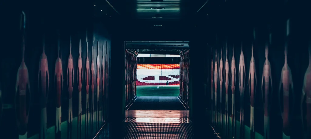 A ground-level view of a professional soccer dugout and technical area, looking out toward the pitch. The scene illustrates a key physical component of the United States soccer pyramid, where coaches and substitute players from various professional tiers&mdash;such as MLS or the USL Championship&mdash;oversee the match from the sidelines.
