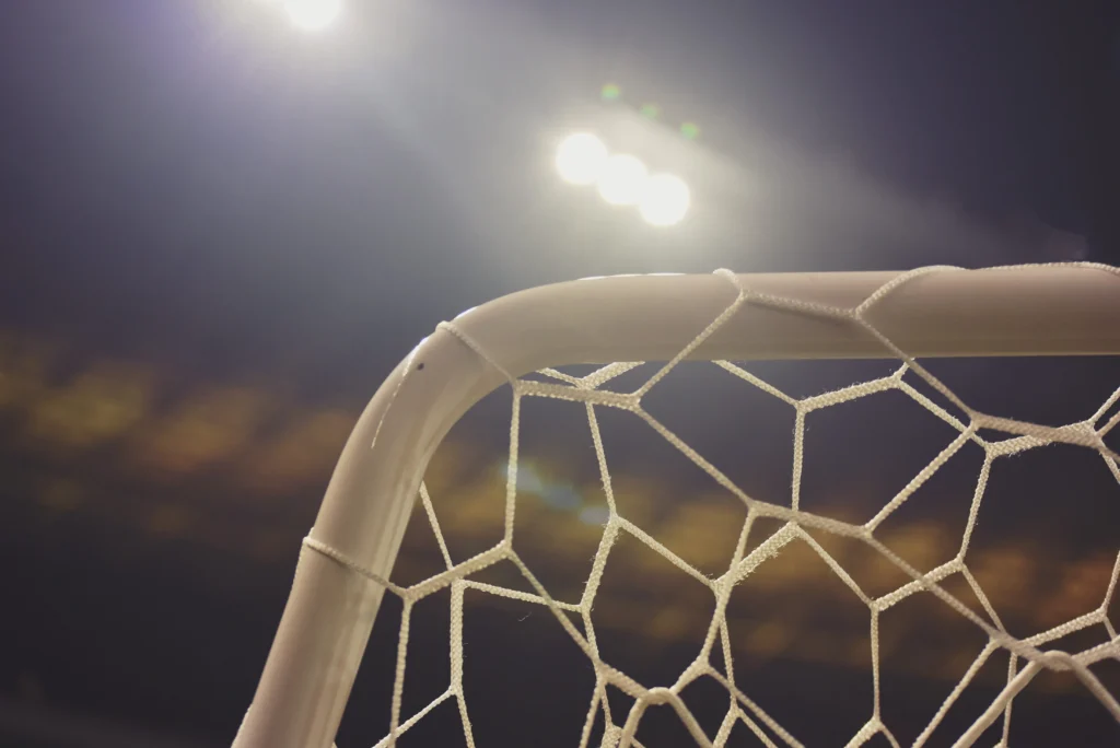 A close-up, artistic shot of the top corner of a white goal post and netting against a dark night sky. The stadium floodlights create a bright glow above the crossbar, highlighting the structure and raising the question: how tall is a football goal?