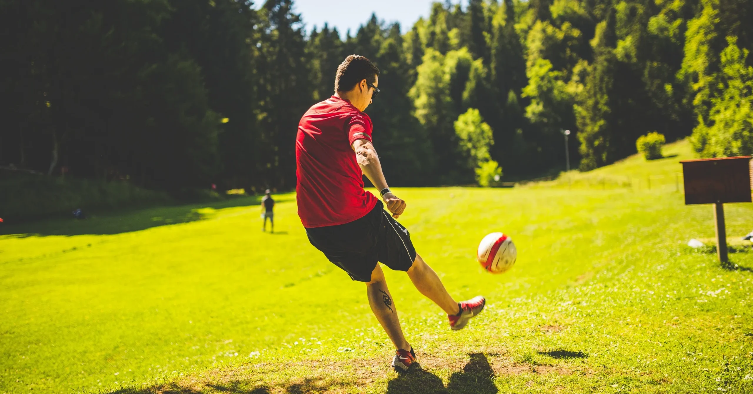 A person in a red shirt and black shorts kicks a ball across a large, open green field lined with trees. This illustrates a typical scene from a game of footgolf, which combines elements of football and golf.