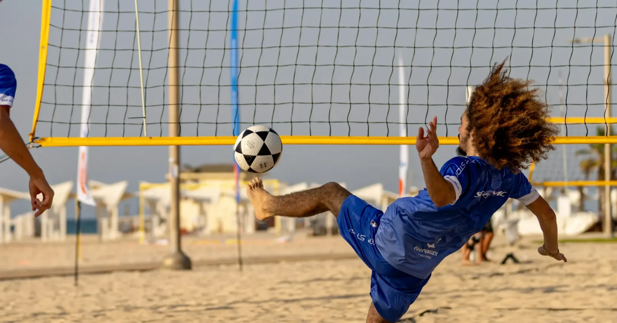 A man with curly hair performs an athletic mid-air kick during a game of footvolley on a sandy beach. He is wearing a blue kit and is positioned in front of a yellow net, with the sun low in the sky behind him.
