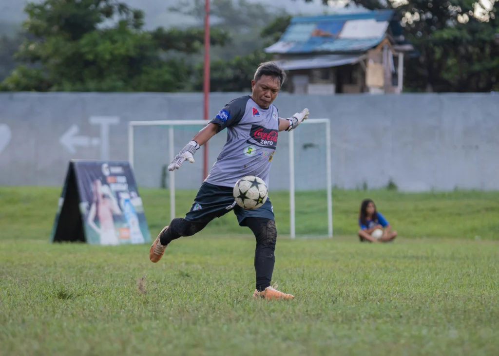 A good goalkeeper in a grey and black kit kicks a football from the grass in an open field, demonstrating precise distribution and technique.