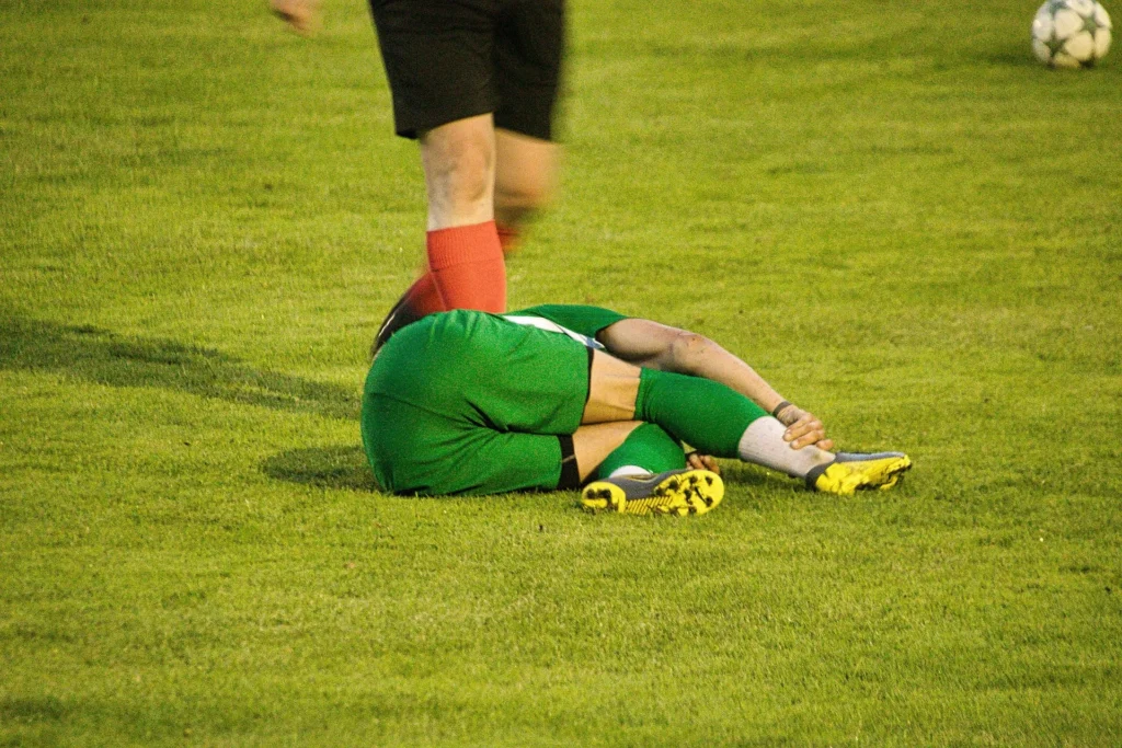 A football player in a green uniform lies on the grass holding their leg after a collision, while another player and the ball are visible nearby during a match. In football, “diving” means a player deliberately falls or exaggerates contact to try to trick the referee into awarding a foul or penalty, but this image instead appears to show a genuine injury during play.