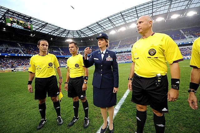 A woman in a dark blue military dress uniform tosses a coin in the air while standing at the center of a professional football pitch. She is flanked by three referees in yellow and black uniforms as they prepare for a high-level football match in a large, crowded stadium.