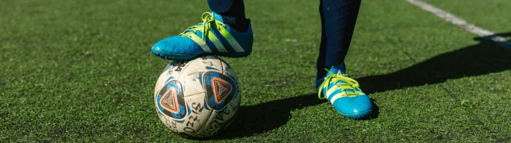 A close-up of a player's legs and blue and yellow footwear on a synthetic turf field, with one foot resting on a ball. This type of footwear is commonly used in both traditional football and footgolf.