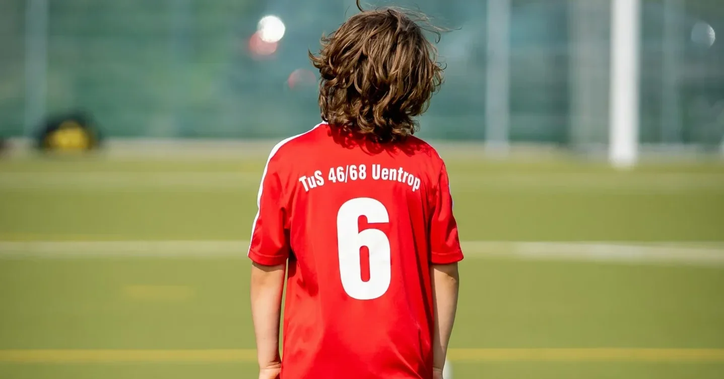 Young soccer player standing on a football pitch wearing a red jersey with the text “TuS 46/68 Uentrop” and the football number 6 on the back, facing the goal in the distance.