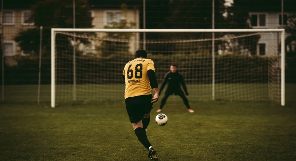 A football player wearing a yellow jersey with the number 68 and "KORKMAZ" on the back takes a penalty kick on a grass field. The player is captured mid-strike, with the ball in the air heading toward the goal. A goalkeeper in black stands centered in the white goal frame, prepared to make a save.
