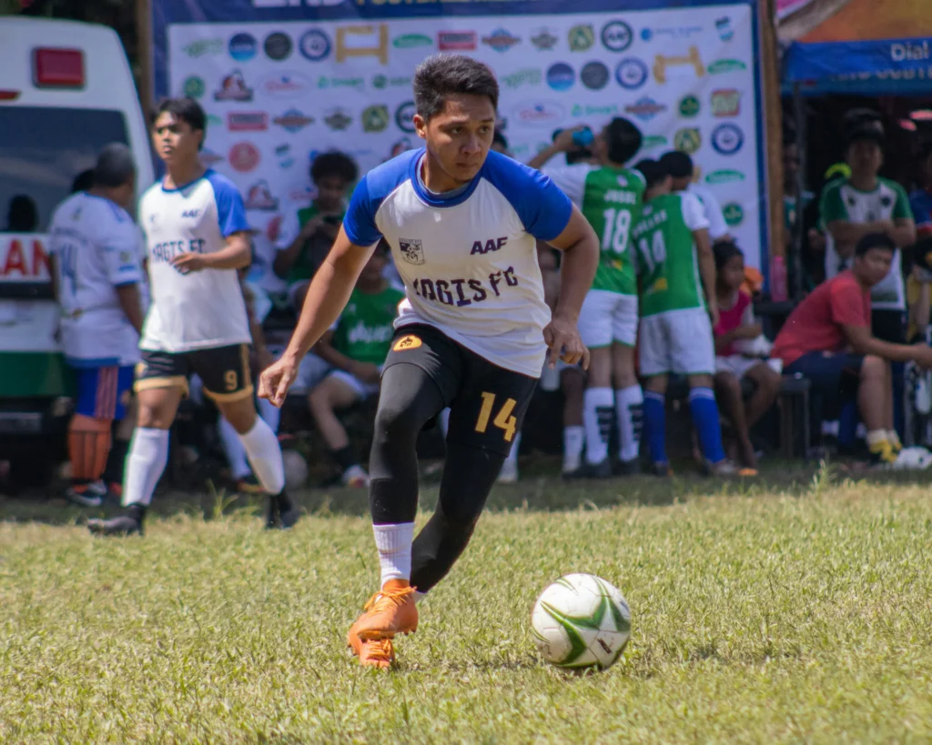A football player wearing a white and blue jersey with number 14 sprinting toward a ball on a grassy pitch during a daytime match, with teammates and spectators behind him, useful for explaining how is possession calculated in football.