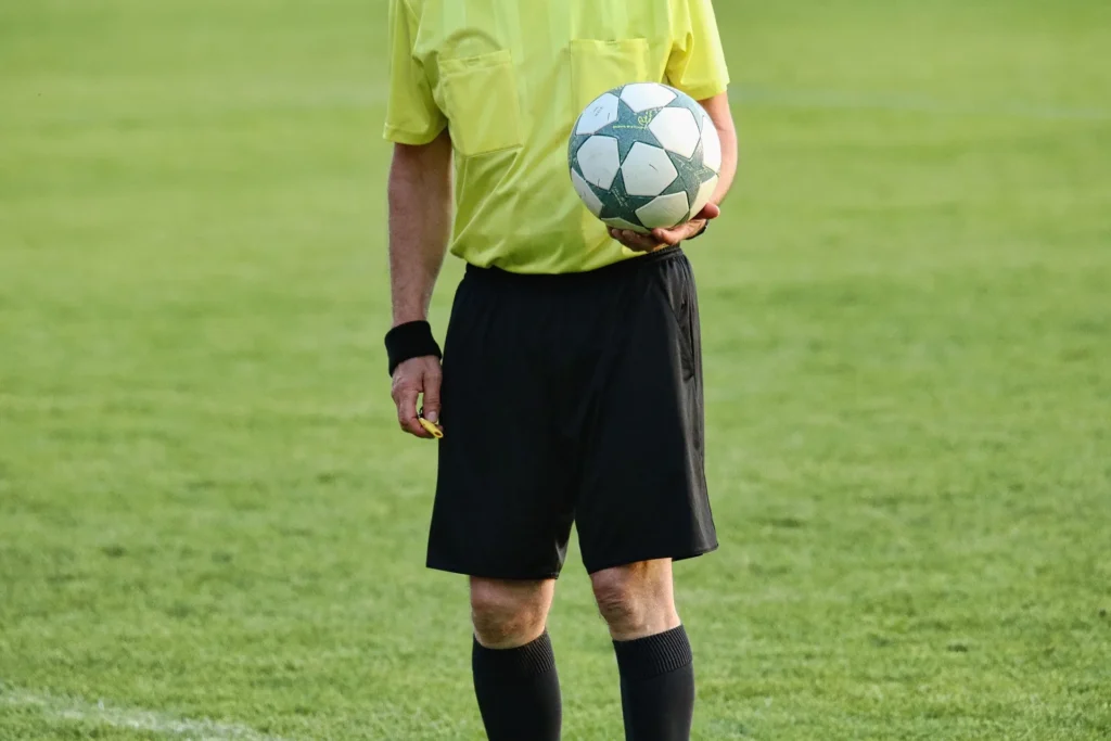 A football referee wearing a yellow shirt and black shorts stands on the pitch holding a match ball and a whistle, ready to officiate the game. Referees watch for rule violations such as fouls and diving in football, which is when a player intentionally falls or pretends to be fouled to gain an unfair advantage.