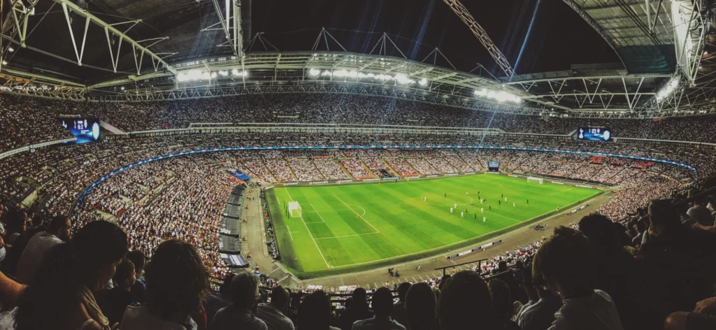 A panoramic internal view of a modern, nearly empty football stadium during the day. The vast rows of seating and the large scale of the architecture provide a sense of the immense capacity found at the biggest football stadium in uk, Wembley Stadium, which seats 90,000 spectators.