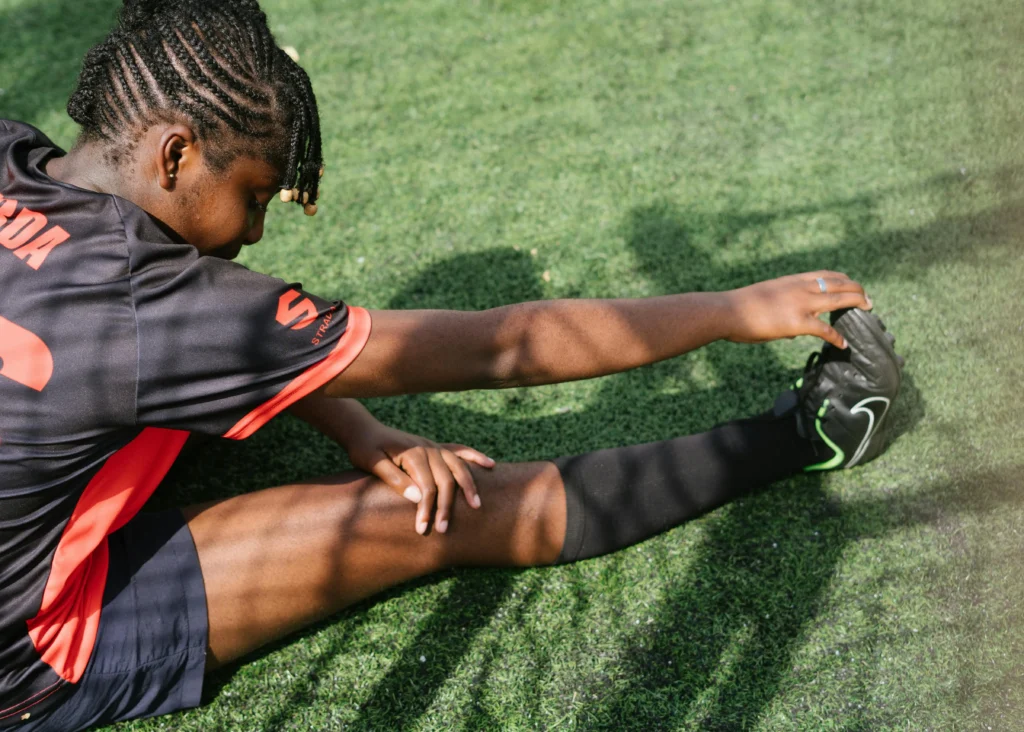 A close-up, high-angle shot of a football player sitting on a green turf field performing stretches for football. The player is wearing a black and red jersey and is reaching forward to pull the toe of their football boot, focusing on a deep hamstring and calf stretch.