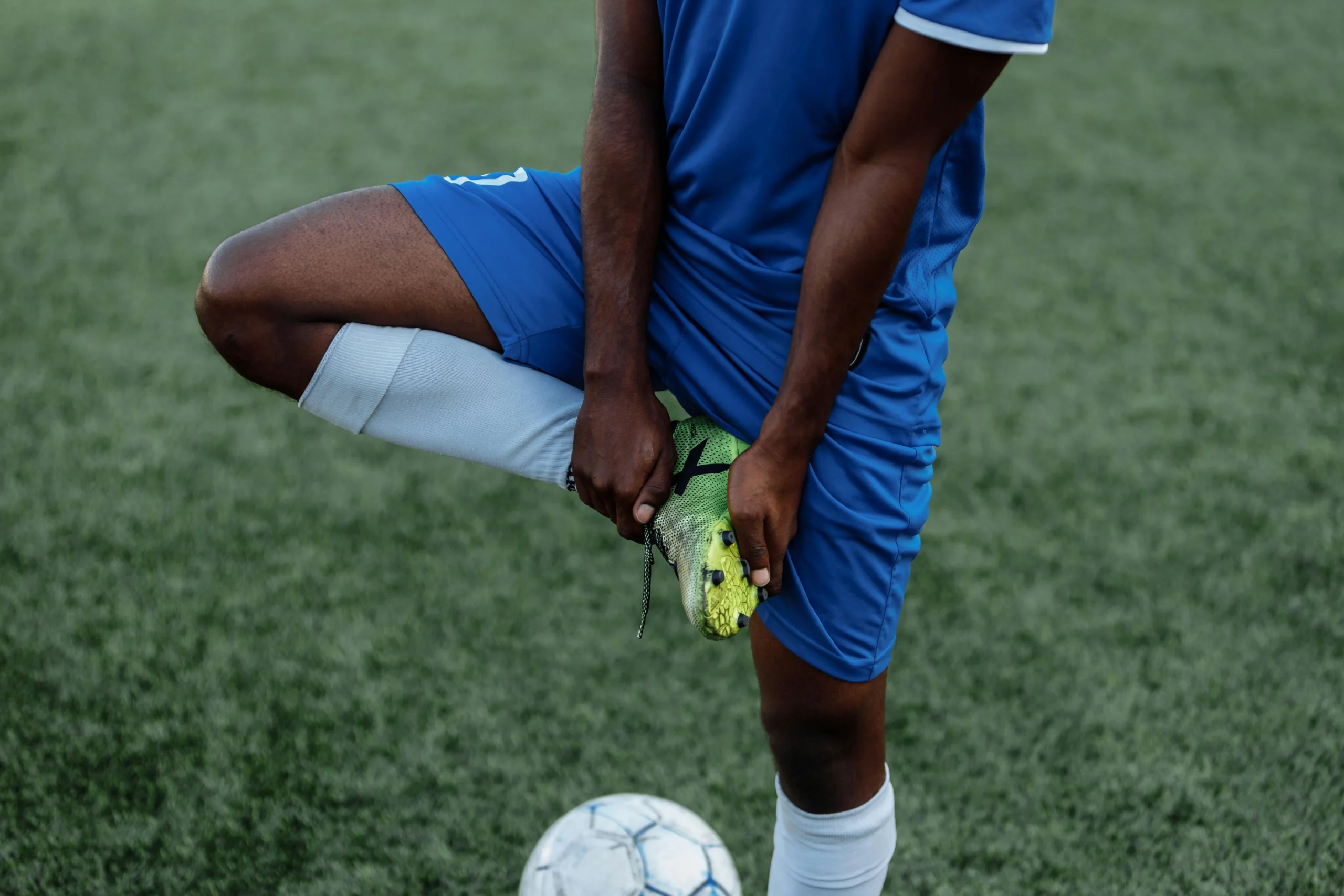 A close-up of a football player in a blue kit performing a standing quadriceps stretch on a turf pitch. This prep-work is a vital component of managing the sprint load in football, ensuring muscle elasticity and joint mobility to prevent injuries during high-speed exertion.