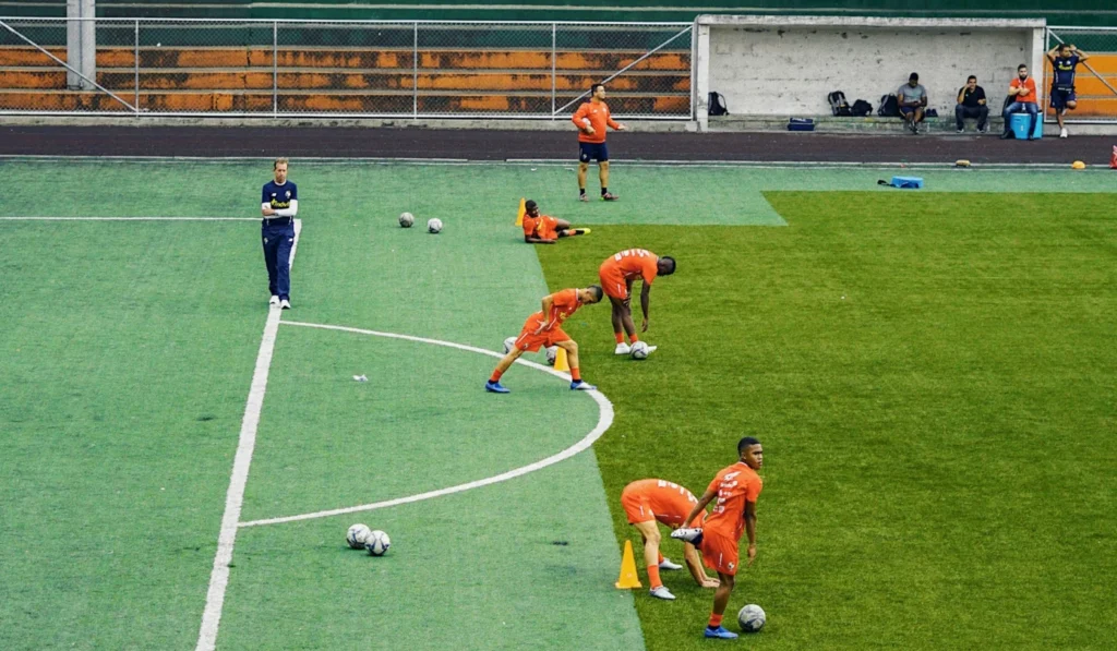 A wide, vertical shot of a football team in bright orange training kits performing various stretches for football across a green artificial turf pitch. In the foreground, two players perform standing quad stretches, while others in the background are seen lunging and bending over to warm up near yellow training cones and footballs.