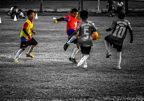 A group of young players in bright jerseys competes for the ball on a grass field in a selective-color image. The intense game-state reading on display highlights the type of advanced decision-making skills scouted during early recruitment in youth football.