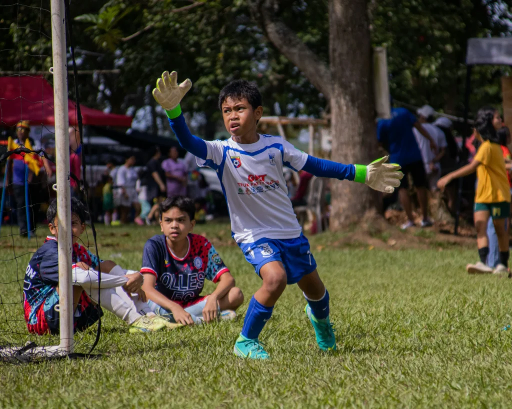 How to be a good goalkeeper? A young player acting as a good goalkeeper stands alert in front of a net with arms wide, wearing green gloves and a white and blue jersey during an outdoor match.