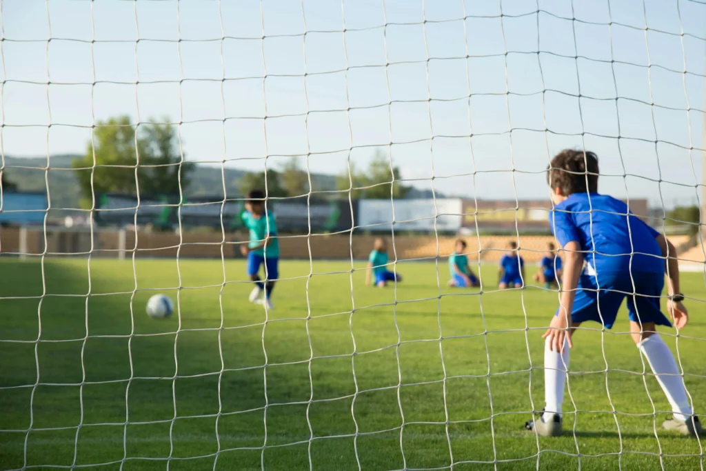 A view from behind a white goal net during a youth football match. In the foreground, a young goalkeeper in a blue kit stands ready in a crouched position. In the blurred background, a player in a teal jersey has just kicked the ball toward the goal, while other teammates watch from the grass.