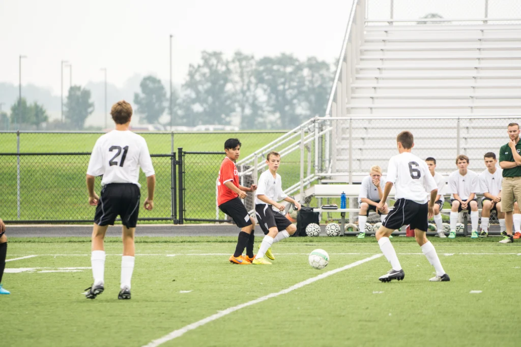 Youth football players in white and red kits compete on a synthetic turf field while coaches and substitutes watch from the sidelines. This competitive environment is where scouts evaluate the cognitive architecture of prospects during the process of early recruitment in youth football.